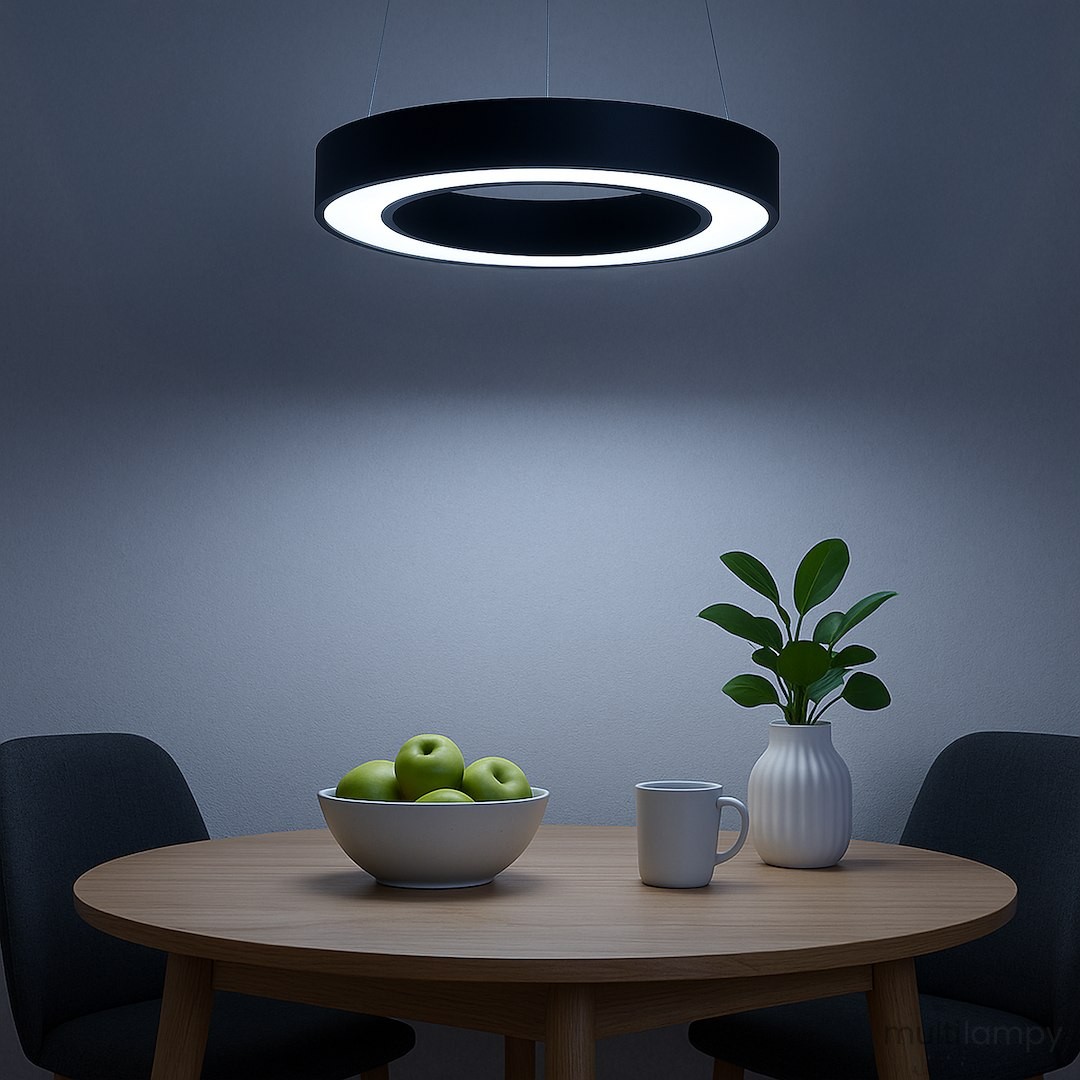Lampa wisząca LED Union 40W CCT w kolorze czarnym, okrągła, z podświetleniem. Zawieszona nad drewnianym stołem z białą misą z zielonymi jabłkami oraz białym kubkiem i doniczką z rośliną. Tło w odcieniach szaro-niebieskich.