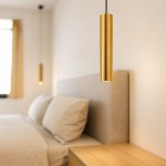 Lampa wisząca Genesis 1xGU10 w kolorze czarnym i złotym, o cylindrycznym kształcie, zawieszona nad łóżkiem w sypialni. W tle widoczne poduszki oraz drewniana szafka nocna. Tło jasne, minimalistyczne.