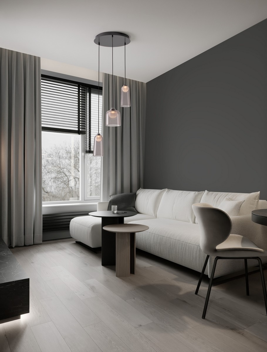 Lampa wisząca Tavenco 3xLED 15W w kolorze czarnym, z trzema szklanymi kloszami, zawieszona na cienkich linkach. Produkt umieszczony w nowoczesnym wnętrzu z jasną sofą i stolikami. Tło w szarościach, z oknem i zasłonami.