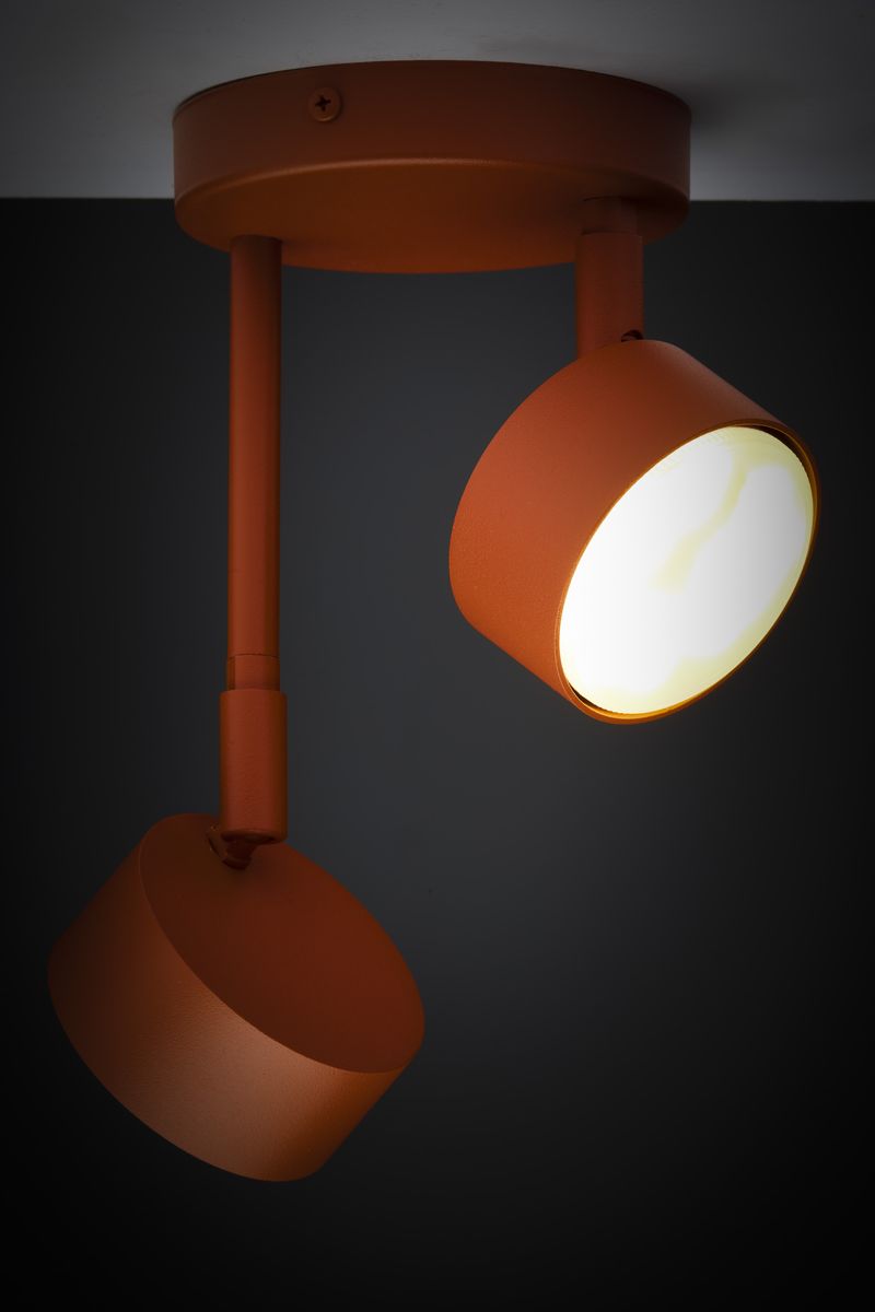 Lampa wisząca Nemo 2xGX53, w kolorze ceglanym, z dwoma cylindrycznymi kloszami. Ujęcie z boku, ukazujące mocowanie sufitowe oraz regulowane ramiona lampy. Klosze mają matową fakturę, emitując ciepłe światło na tle ciemnej ściany.