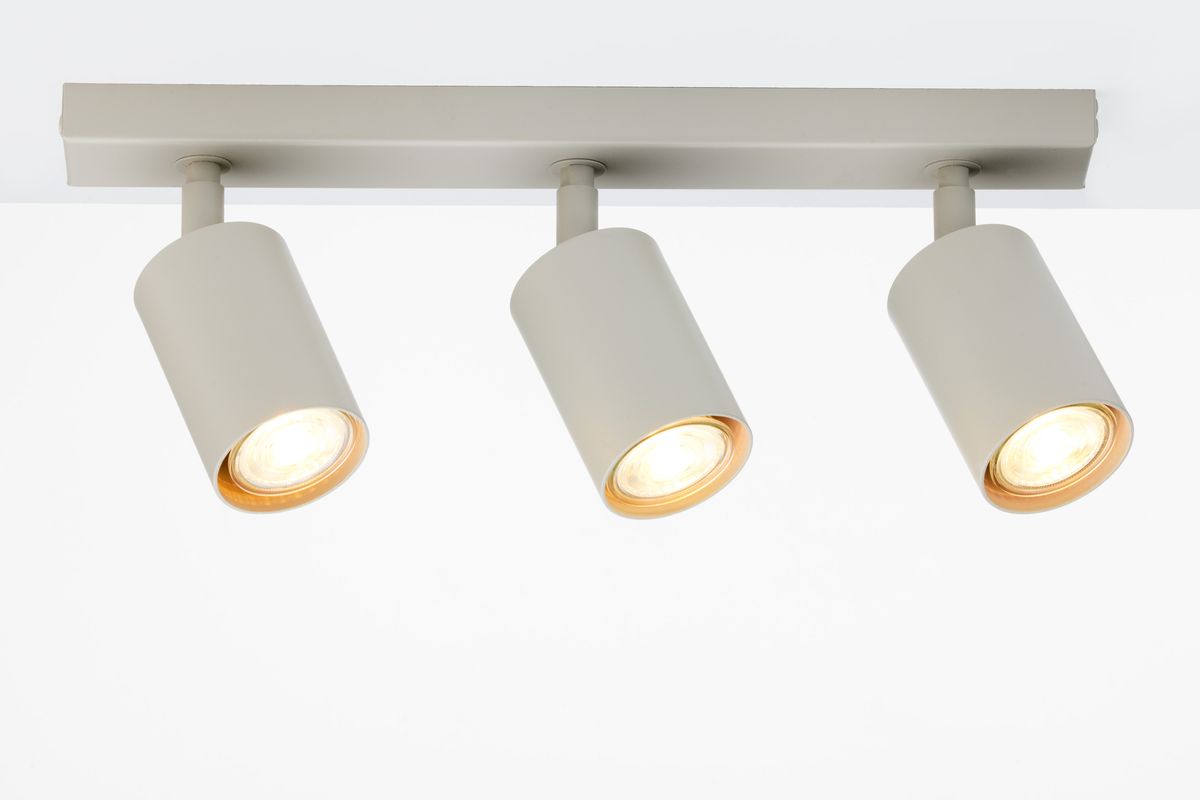 Lampa sufitowa Zoom z trzema spotami GU10 w odcieniu szarym beżowym. Ułożona poziomo, z cylindrycznymi kloszami skierowanymi w dół. Metalowa konstrukcja, minimalistyczny design, widoczne ciepłe światło z żarówek.