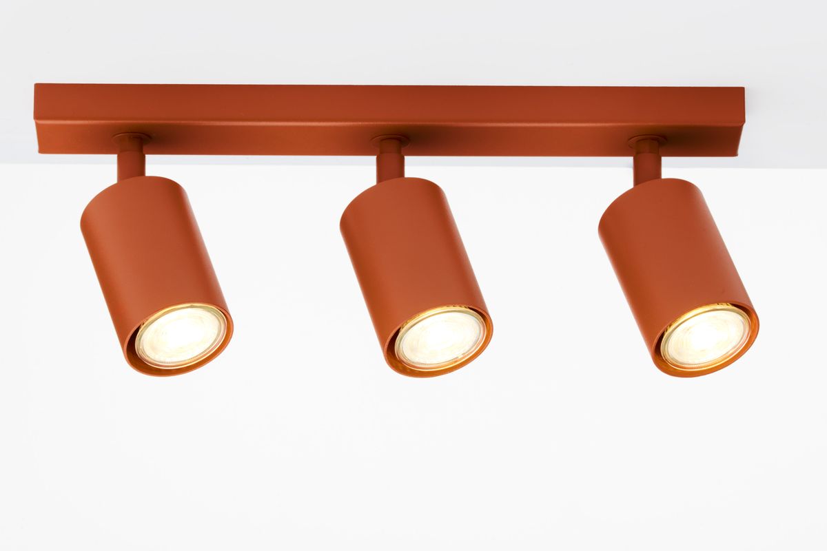 Lampa sufitowa Zoom z trzema reflektorami GU10 w ceglastym kolorze. Ułożona poziomo, z cylindrycznymi kloszami skierowanymi w dół, zamocowana na prostokątnej podstawie. Idealna do oświetlenia wnętrz.