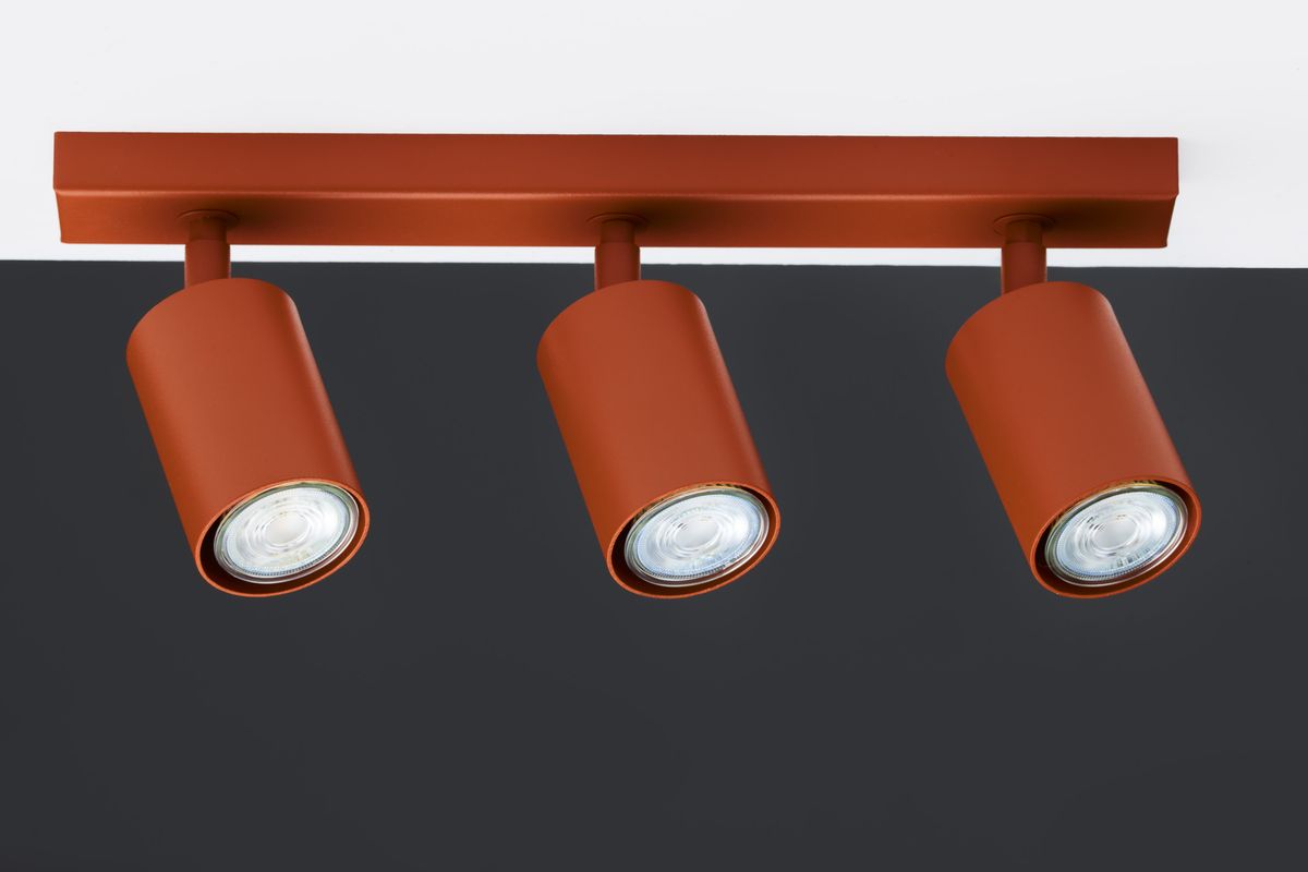 Lampa sufitowa Zoom w kolorze ceglastym z trzema reflektorami GU10. Ujęcie frontalne, pokazujące prostokątny stelaż oraz cylindryczne oprawy. Wykończenie matowe, nowoczesny design. Tło w ciemnym kolorze, podkreślające lampę.