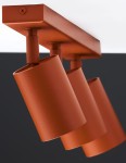 Lampa sufitowa Zoom z trzema reflektorami GU10 w ceglastym kolorze. Reflektory mają cylindryczny kształt, są zamocowane na prostokątnej podstawie. Ujęcie z boku, ukazujące detale konstrukcji i teksturę powierzchni.