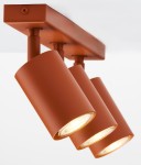 Lampa sufitowa Zoom z trzema reflektorami GU10 w ceglastym kolorze. Reflektory mają cylindryczny kształt, zamontowane na prostokątnej podstawie. Ujęcie z dołu, pod kątem, ukazujące szczegóły konstrukcji i źródła światła.