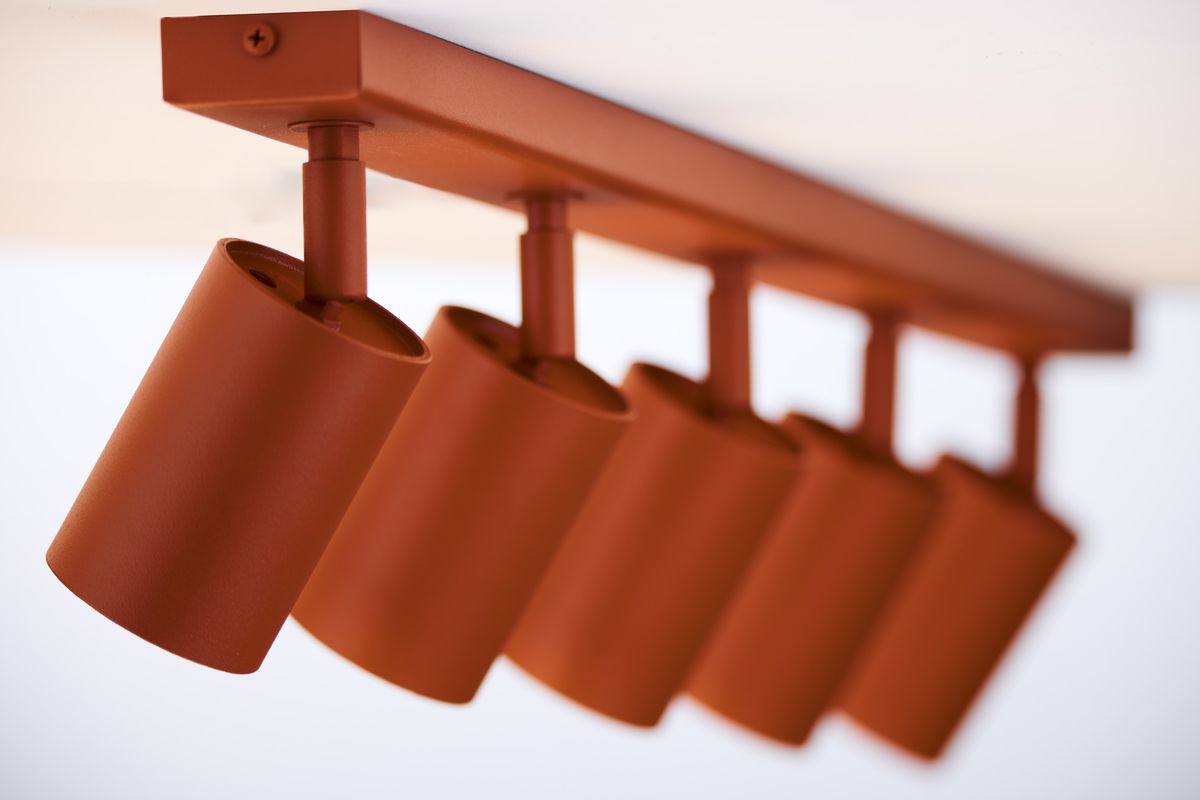 Lampa sufitowa typu spot w ceglastym kolorze z pięcioma oprawami GU10. Ujęcie z boku, ukazujące cylindryczne klosze zawieszone na prostokątnej podstawie. Matowa faktura metalu, stabilnie zamontowana pod sufitem.