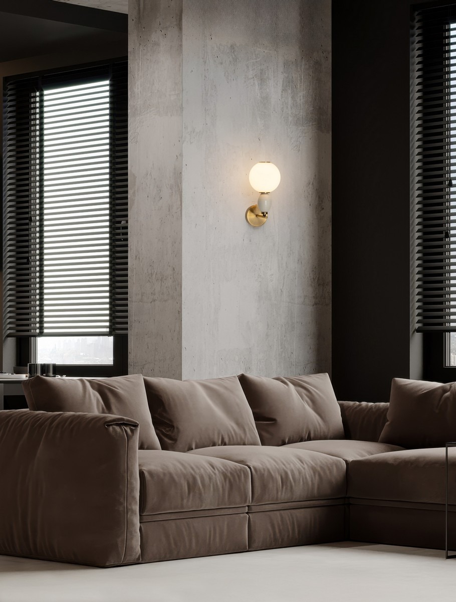 Laverzo kinkiet 1xLED 6W w kolorze złotym i beżowym, zamontowany na ścianie, z białym, okrągłym kloszem. W tle widoczna brązowa sofa i okna z żaluzjami. Kinkiet o minimalistycznym designie, zapewniający ciepłe światło.