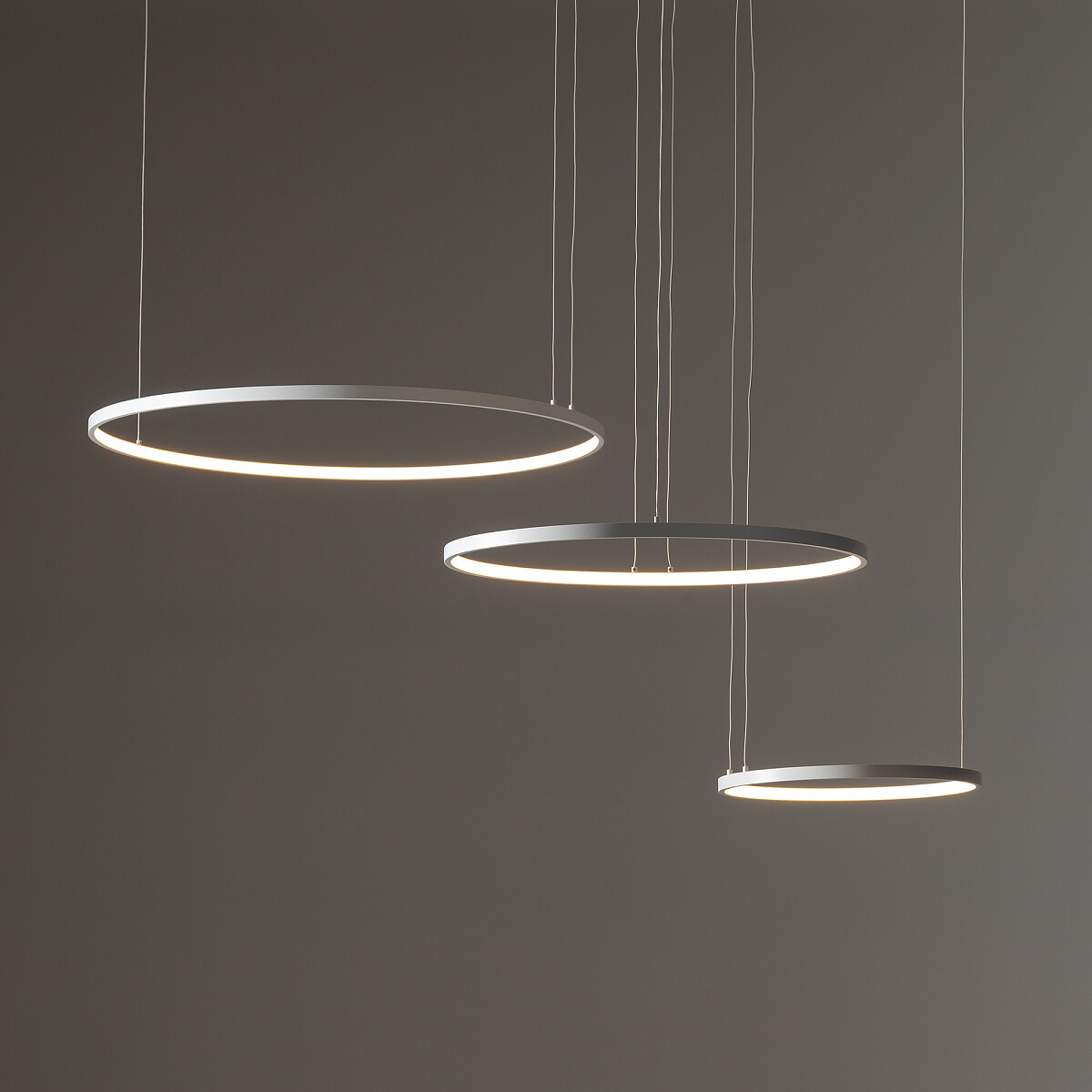 Lampa wisząca LED Circolo 85W, biała, z trzema okrągłymi, metalowymi pierścieniami o różnej średnicy, zawieszona na cienkich linkach. Źródło światła o temperaturze 3000K, idealna do nowoczesnych wnętrz. Tło w odcieniach szarości.