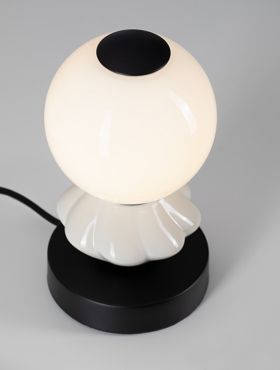 Lampa stołowa Monreta z jedną żarówką LED 6W, o okrągłej, mlecznej kloszu i czarnej, matowej podstawie. Klosz umieszczony na dekoracyjnej, jasnoszarej podstawie w kształcie kwiatu. Produkt widoczny z góry, na jednolitym tle.