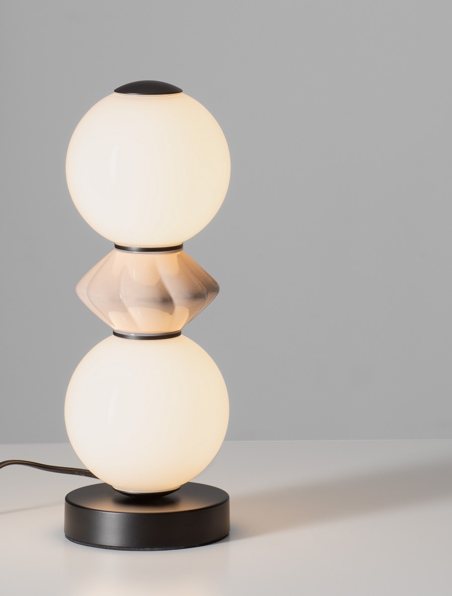 Lampa stołowa Monreta z czarną podstawą, składająca się z dwóch okrągłych kloszy w kolorze białym oraz jednego dekoracyjnego elementu w odcieniu mokka. Ujęcie frontalne, produkt stoi na płaskiej powierzchni.