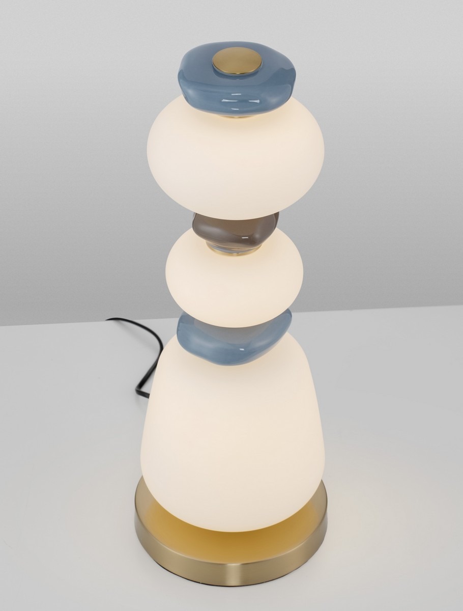 Lampa stołowa Felanzo z jedną żarówką LED 23W. Okrągła, biała szklana podstawa z dekoracyjnymi, kolorowymi elementami (niebieskim i brązowym). Metalowa, złota podstawa. Ujęcie z góry, na białym tle.