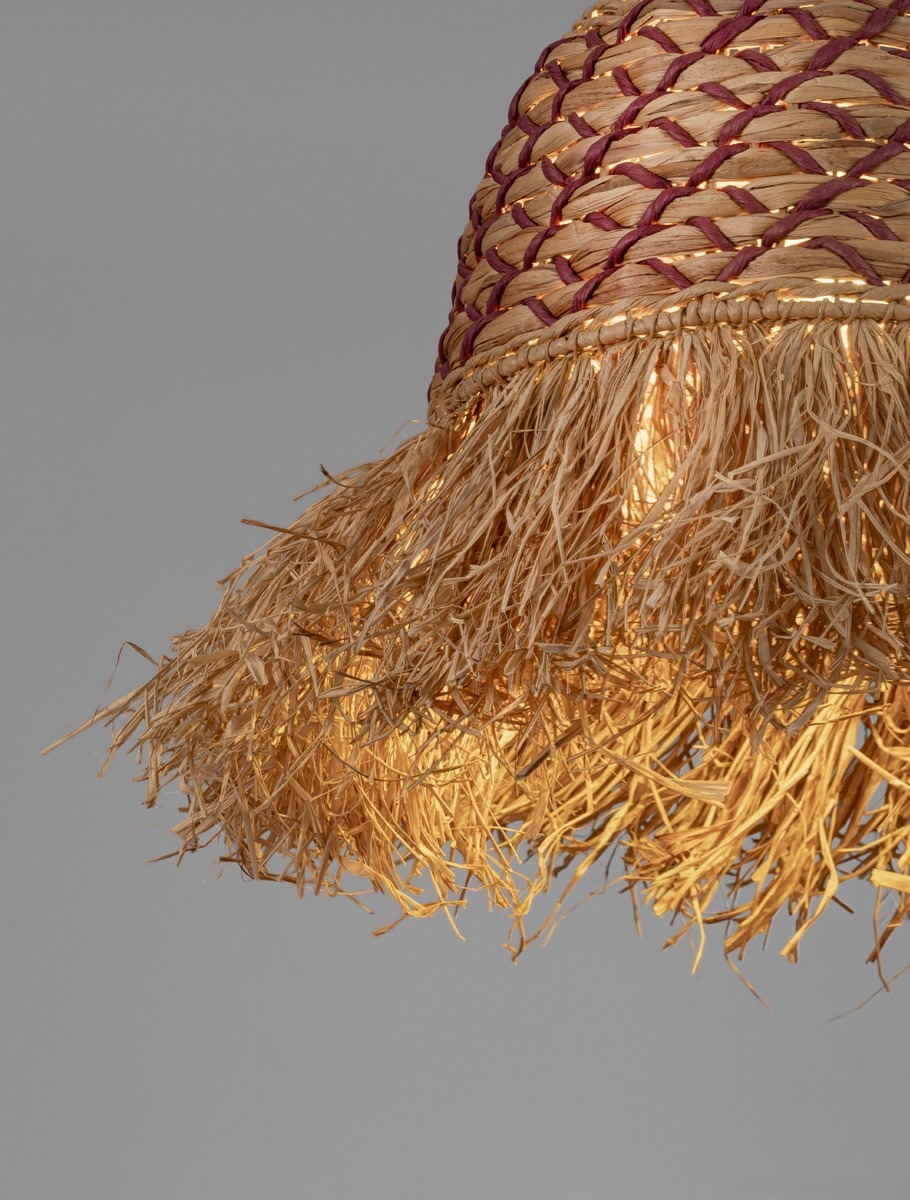 Lampa wisząca Fanliza z kloszem w kolorze naturalnym, z plecionką, wykończona frędzlami z trawy. Klosz ma okrągły kształt, podwieszony, z miejscem na żarówkę E27. Tło szare, minimalistyczne, podkreślające detal lampy.