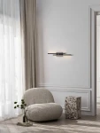 Kinkiet Felvira 2xLED 12W w kolorze czarnym, zamontowany na ścianie w minimalistycznym wnętrzu. Obok widoczny miękki fotel i dekoracyjny stolik. Światło emitowane przez kinkiet subtelnie oświetla przestrzeń.