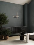 Kinkiet Catervo 2xLED 14W w kolorze czarnym, z minimalistycznym wzornictwem. Zainstalowany na ciemnej ścianie, składa się z poziomego i pionowego elementu w odcieniu złotym. W tle widoczne rośliny oraz nowoczesne meble.