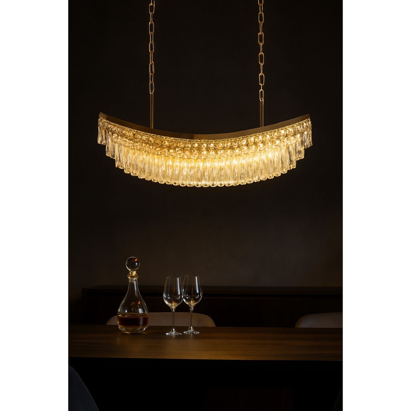 Lampa wisząca Zambezi w kolorze złotym, o półokrągłym kształcie, z 18 miejscami na żarówki E14. Posiada dekoracyjne, przezroczyste kryształki zwisające na dolnej krawędzi. Zawieszona na dwóch łańcuchach w ciemnym otoczeniu, nad drewnianym stołem.