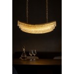 Lampa wisząca Zambezi w kolorze złotym, o półokrągłym kształcie, z 18 miejscami na żarówki E14. Posiada dekoracyjne, przezroczyste kryształki zwisające na dolnej krawędzi. Zawieszona na dwóch łańcuchach w ciemnym otoczeniu, nad drewnianym stołem.