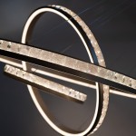 Lampa wisząca Camelia z trzema źródłami LED, w złotym kolorze. Składa się z dwóch okrągłych pierścieni, zdobionych przezroczystymi kryształkami, emitujących ciepłe światło. Zawieszona na cienkich linkach, ma nowoczesny, minimalistyczny design.