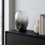Lampa stołowa LED Pamela 12W w czarnym kolorze, z transparentnym kloszem o zaokrąglonym kształcie, umieszczona na czarnej powierzchni. W tle widoczne książki i biały wazon. Produkt stoi na poziomie blatu, w jasnym wnętrzu.
