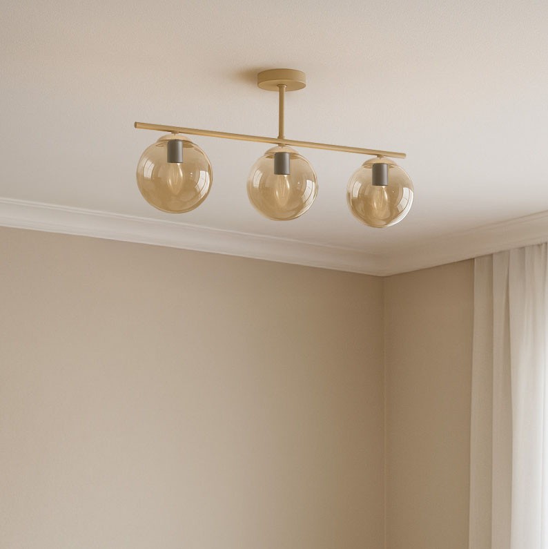 Lampa sufitowa Felix z trzema kloszami w kształcie kul, wykonana z bursztynowego szkła, zawieszona na beżowej metalowej ramie. Umieszczona na suficie w jasnym wnętrzu, z delikatnym tłem w odcieniach beżu.