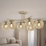 Beżowa lampa sufitowa Felix z sześcioma kloszami w bursztynowym kolorze, o okrągłym kształcie, zamontowana na metalowej ramie. Ujęcie z dołu, pokazujące detal świecących żarówek w szklanych kulach, w tle jasne wnętrze z sofą.