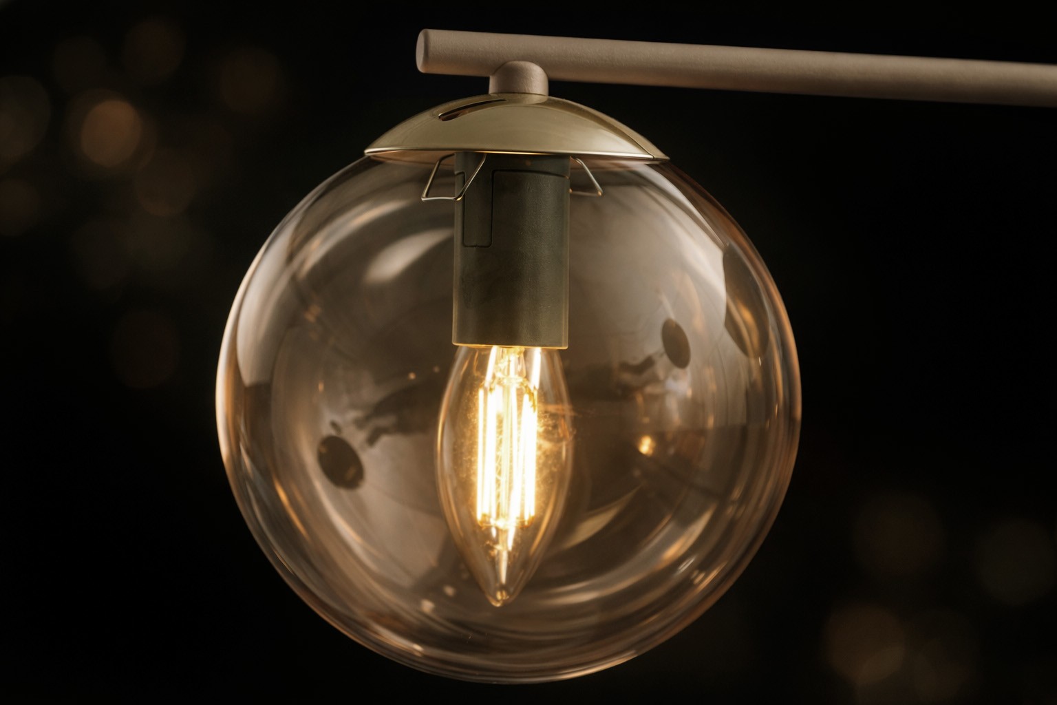 Kinkiet Felix w odcieniach beżowym i bursztynowym, z okrągłą, przezroczystą kloszem. Żarówka E14 w kształcie łzy umieszczona w metalowym mocowaniu. Kinkiet zawieszony na prostym, metalowym ramieniu, na ciemnym tle.