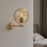 Kinkiet Felix 1xE14 w odcieniu beżowym z bursztynową szklaną kopułą, zamontowany na ścianie. Prosta, okrągła podstawa oraz wygięty uchwyt. Widoczna żarówka w środku, w tle łóżko z jasną pościelą.