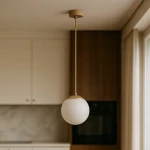 Lampa wisząca Savia z beżowym mocowaniem i białym kloszem w kształcie kuli. Zawieszona na cienkim przewodzie, idealna do nowoczesnych wnętrz. Widoczna w kuchni, na tle drewnianej ściany i jasnych mebli.