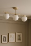Beżowa lampa sufitowa Savia z trzema białymi kloszami w kształcie kul, zamontowana na złotej metalowej rurze, wisząca w pokoju. Tło w neutralnych odcieniach z obrazami w złotych ramach.