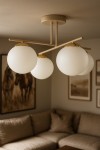 Lampa sufitowa Savia w kolorze beżowym i białym z czterema kulistymi kloszami z matowego szkła. Ujęcie frontalne, lampa zamontowana na suficie, otoczona stonowanym wnętrzem z kanapą i obrazami na ścianach.
