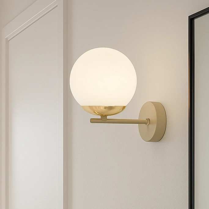 Beżowy kinkiet Savia z oprawą na żarówkę E14, z okrągłym, białym kloszem z matowego szkła i metalowym uchwytem w kolorze złotym. Kinkiet zamontowany na jasnej ścianie, w nowoczesnym stylu.