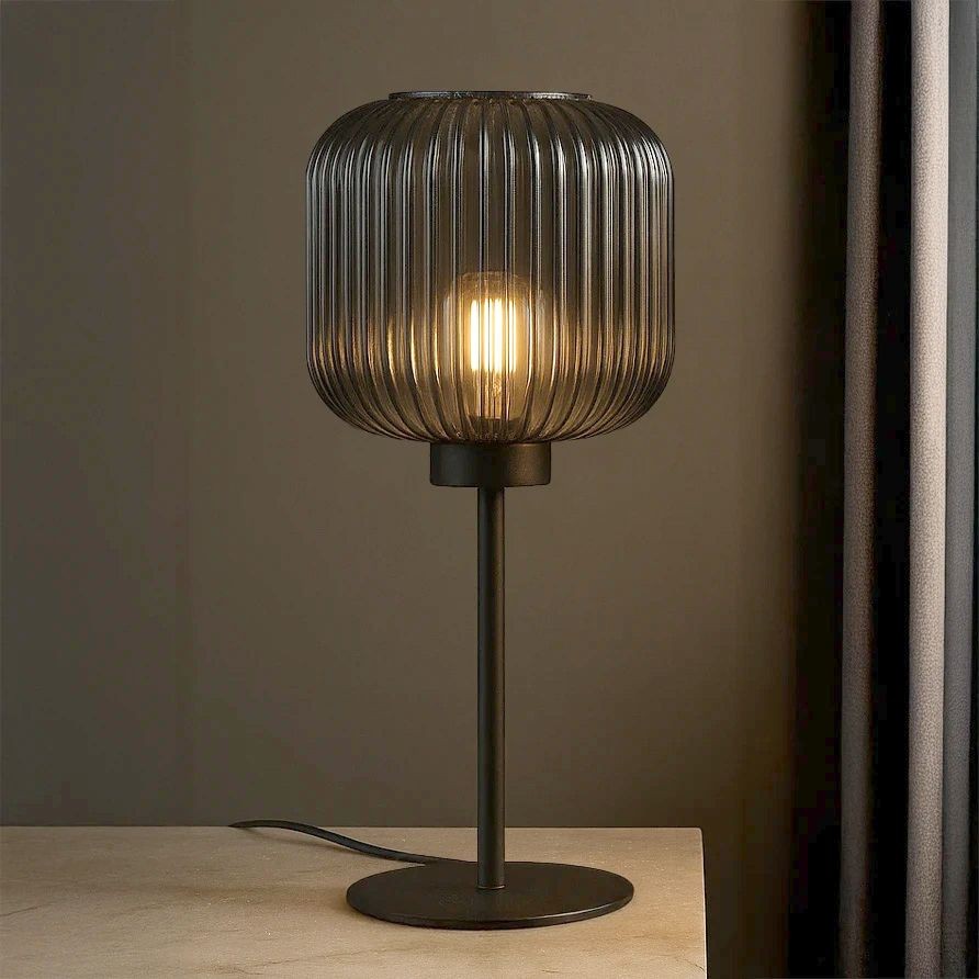 Lampa stołowa Campo z oprawą w kolorze czarnym i grafitowym, z przezroczystym, żłobkowanym kloszem. Stojąca, z okrągłą podstawą, z widoczną żarówką E27, na jasnym tle. Idealna do nowoczesnych wnętrz.