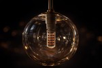 Lampa wisząca Galaxy K-5560 w kolorze czarnym z bursztynowym kloszem. Okrągły, szklany klosz z widoczną żarówką G9. Ujęcie z przodu, zawieszona na metalowej konstrukcji, na ciemnym tle.