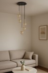 Lampa wisząca Galaxy z pięcioma kulistymi kloszami w bursztynowym kolorze, zawieszona na czarnych przewodach. Metalowe mocowania w kolorze złotym. Umiejscowiona w jasnym wnętrzu, nad beżową sofą i okrągłym stołem.