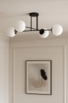Czarna lampa sufitowa Savoy 4-punktowa z białymi okrągłymi kloszami, zamontowana na suficie. Klosze rozmieszczone w linii, podkreślają minimalistyczny design. Tło jasne, z obrazem w ramie na ścianie obok.