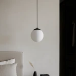Czarna lampa wisząca Savoy D16 z białym, okrągłym kloszem, zawieszona na długim przewodzie. Klosz ma matową powierzchnię, a lampa znajduje się w jasnym wnętrzu, nad łóżkiem z jasną pościelą i dekoracjami.