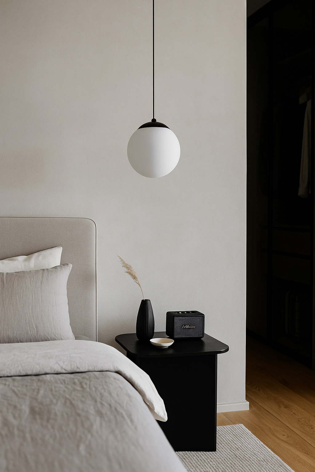 Lampa wisząca Savoy D16 w kolorze czarnym z okrągłym, białym kloszem zawieszona w pomieszczeniu. Wyeksponowana w sypialni nad czarnym stolikiem nocnym, obok dekoracyjnego wazonu i radia. Tło w odcieniach szarości.