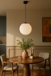 Lampa wisząca Savoy D30 w kolorze czarnym z okrągłym, mlecznym kloszem, zawieszona na cienkim przewodzie. Ujęcie frontalne, z lampą w centralnej części nad okrągłym drewnianym stołem, w otoczeniu dekoracyjnych elementów.