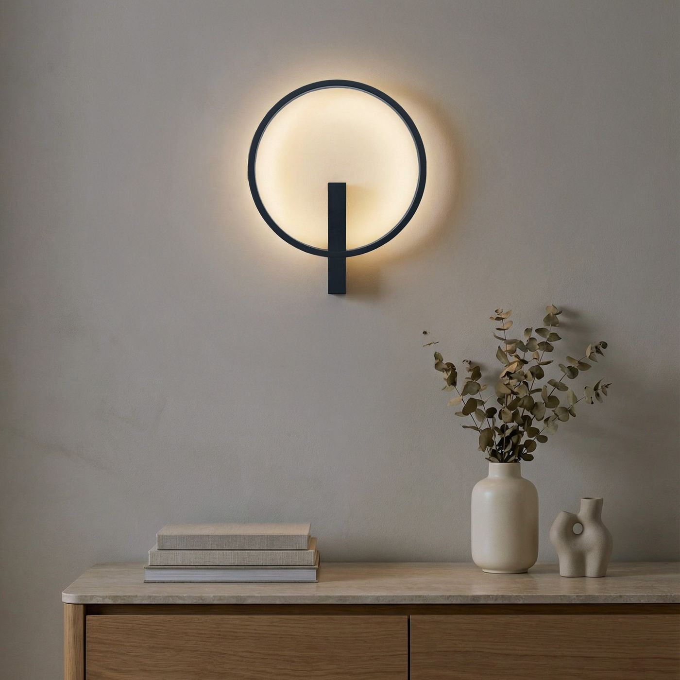Kinkiet LED Arte o mocy 25W w kolorze czarnym, w kształcie okręgu, montowany na ścianie. Emituje ciepłe światło, z widoczną metalową podstawą. W tle dekoracyjny stół z książkami oraz wazonem z suszonymi gałązkami.