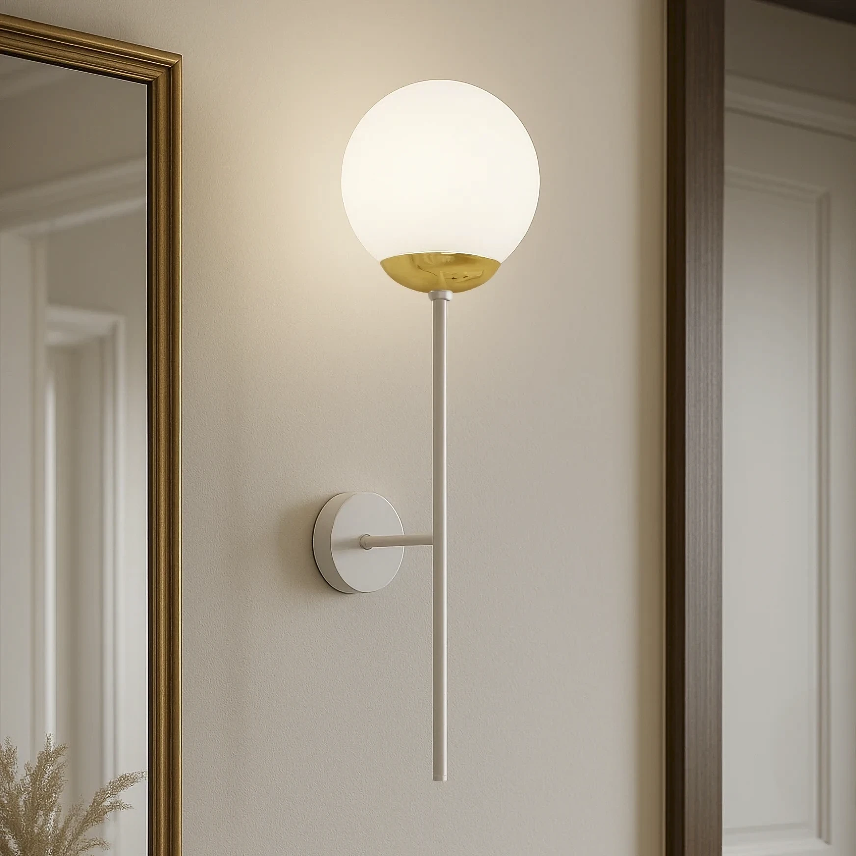 Kinkiet Elmo 1xE14 w kolorze białym i złotym, z okrągłą, półprzezroczystą kloszem, zamontowany na ścianie. Klosz w górnej części złoty, w dolnej mleczny. Prosta, metalowa konstrukcja w formie pionowego ramienia. W tle lustro w złotej ramie.