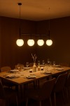 Lampa wisząca 4-punktowa Faro w czarnym kolorze z czterema okrągłymi kloszami w odcieniu ciepłej bieli. Zawieszona równolegle nad drewnianym stołem w eleganckiej jadalni, z ustawionymi talerzami i szklankami. Tło w stonowanych brązach.