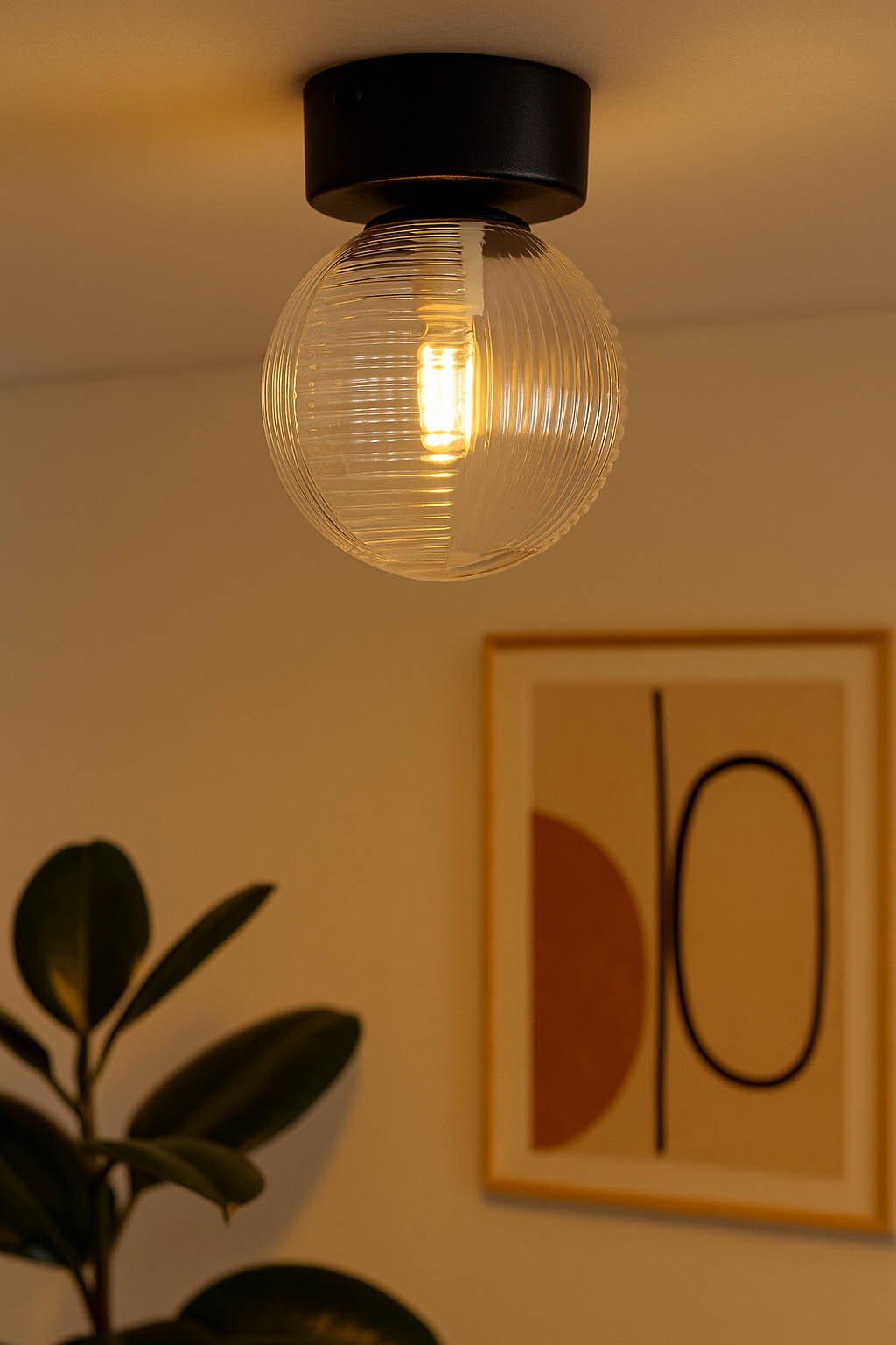 Lampa sufitowa Aura z czarnym stelażem i transparentną, szklaną kulą. Widok z dołu, zawieszona na suficie, z widoczną żarówką G9. W tle roślina doniczkowa oraz abstrakcyjny obraz na ścianie.