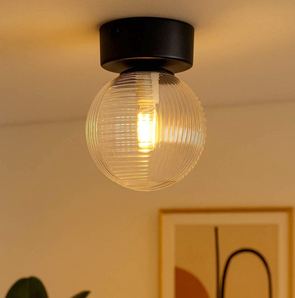 Lampa sufitowa Aura z pojedynczym gniazdem G9, w kolorze czarnym z przezroczystą, teksturowaną kulą. Ujęcie z dołu, ukazujące detal źródła światła w szklanej oprawie. Tło w neutralnych odcieniach, z widocznym fragmentem dekoracji ściennej.