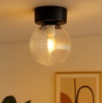 Lampa sufitowa Aura z pojedynczym gniazdem G9, w kolorze czarnym z przezroczystą, teksturowaną kulą. Ujęcie z dołu, ukazujące detal źródła światła w szklanej oprawie. Tło w neutralnych odcieniach, z widocznym fragmentem dekoracji ściennej.