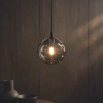 Lampa wisząca Dori z jedną żarówką G9, w kolorze czarnym z grafitowym kloszem. Owalny, przezroczysty klosz z lekkim przydymieniem, zawieszony na cienkim kablu, emitujący ciepłe światło. W tle drewniana ściana oraz ciemne naczynia.