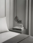 Lampa wisząca Dori w kolorze czarnym i grafitowym z przezroczystą kulistą osłoną, zawieszona nad nocnym stolikiem. Tło w odcieniach szarości z pościelą i książkami, w aranżacji sypialni.
