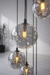Lampa wisząca Dori, czarna z grafitowymi detalami, z osiemnastoma przezroczystymi kloszami w kształcie kul, zawieszona na cienkich przewodach. W tle rośnie roślina w doniczce na jasnym tle.