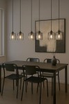 Lampa wisząca Miri, sześć kloszy w kolorze czarnym i grafitowym, zawieszona nad stołem. Klosze mają przezroczyste szkło, a lampa jest zawieszona na długich linkach. W tle widoczny obraz oraz czarny stół z krzesłami.