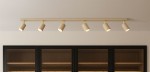 Mati lampa sufitowa beżowa z sześcioma reflektorami GU10, zamontowana na poziomej listwie. Reflektory mają zaokrąglony kształt i są skierowane w dół, umieszczone na jasnym tle nad czarnymi szafkami z drewnianymi półkami.