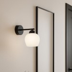 Kinkiet Abro 1xE27 w kolorze czarnym i białym, z okrągłą, matową szklaną kloszem, zamontowany na ścianie. Produkt ma prostą, metalową konstrukcję i jest umieszczony obok ramy na ścianie w jasnym wnętrzu.