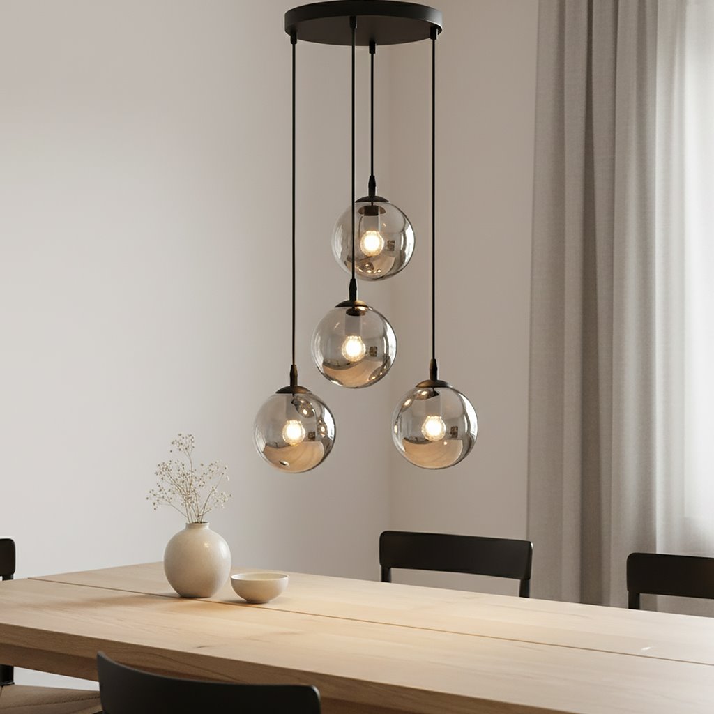 Lampa wisząca Cosmo Premium 4-punktowa w kolorze grafitowym, zawieszona na trzech smukłych linkach. Posiada cztery szklane klosze w kształcie kul, emitujące ciepłe światło, umieszczone nad drewnianym stołem w jasnym wnętrzu.